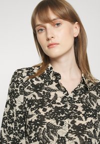 Blusa de botão floral com fundo creme, apresentando padrões de samambaias pretas, um colarinho e um corte solto. Tecido suave com uma textura lisa.