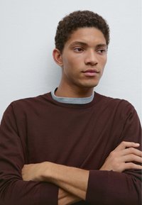 Modello che indossa una camicia a manica lunga color borgogna con un colletto grigio. Ha i capelli ricci e le braccia incrociate, su uno sfondo semplice.
