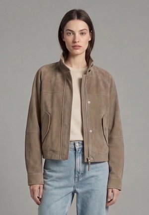 Kvinde iført en taupe ruskind bomberjakke over en beige sweater og lyseblå højtaljede jeans, stående mod en ensfarvet grå baggrund.