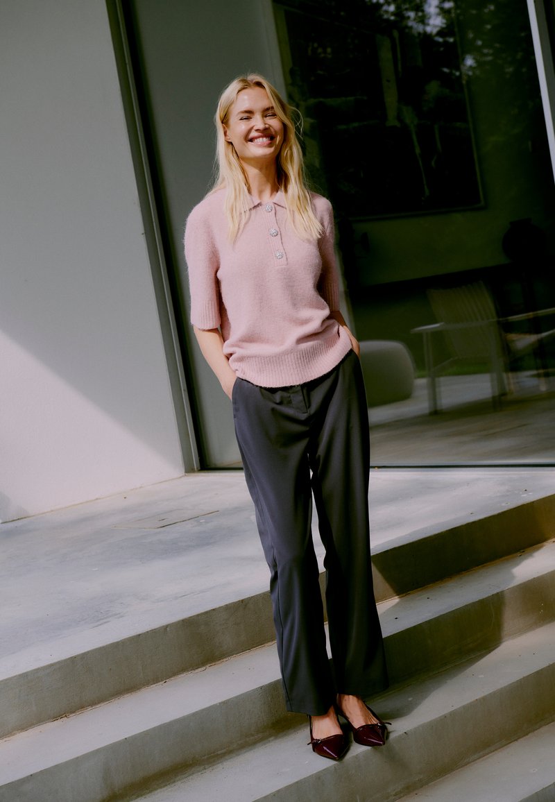 Pull court à manches courtes de couleur rose clair, avec un col et des boutons ; associé à un pantalon ample de couleur sombre et des chaussures bordeaux à bouts pointus.