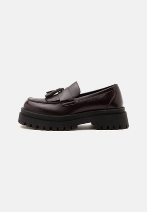 Mocassin en cuir brun foncé avec semelle épaisse noire à crampons et pompons décoratifs sur le dessus, présenté de profil sur fond blanc.