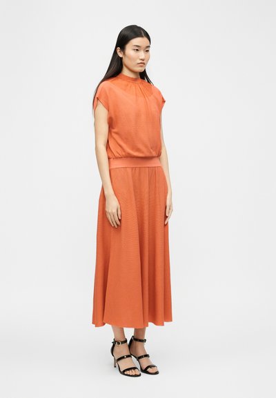 Femme debout portant une robe midi orange sans manches avec une taille froncée et des sandales à talons hauts noires à brides, sur un fond uni.