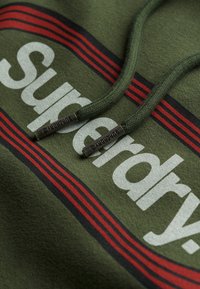 Zielona bluza z dużym białym logo "Superdry", czerwonymi i czarnymi poziomymi paskami oraz plecionymi zielonymi sznurkami z metalowymi końcówkami.