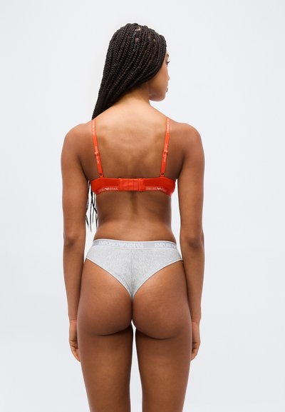 Γυναίκα με πλεξουδάκια που φοράει πορτοκαλί σουτιέν και γκρι εσώρουχα με το λογότυπο Emporio Armani, εμφανιζόμενη από πίσω σε απλό φόντο.