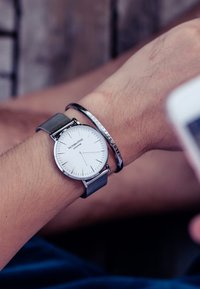 Reloj plateado con una carátula blanca redonda y marcadores de hora plateados, con una correa de malla gris. Lleva una pulsera delgada con texto al lado.