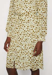 Abito floreale azzurro chiaro con fiori marroni e gialli, dotato di cintura da annodare in vita, maniche lunghe e uno spacco all'altezza del ginocchio. Tessuto dal tocco morbido.