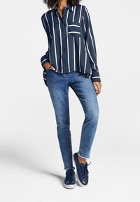 Mujer vistiendo una camisa azul marino de manga larga con botones y rayas verticales blancas, jeans azules con dobladillos deshilachados y deportivas azul marino.