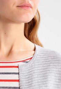 Close-up van het onderste gezicht, de nek en schouder van een vrouw die een grijze geribbelde cardigan draagt over een rood, wit en zwart gestreept shirt.