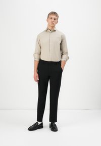 Jonge man die met handen in zijn zakken staat, gekleed in een beige button-up shirt met opgerolde mouwen en zwarte slim-fit broek en instappers.