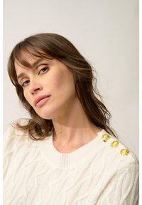 Kremowy sweter o splocie w warkocz z okrągłym dekoltem i trzema złotymi dekoracyjnymi guzikami na lewym ramieniu. Miękka, teksturowana tkanina.