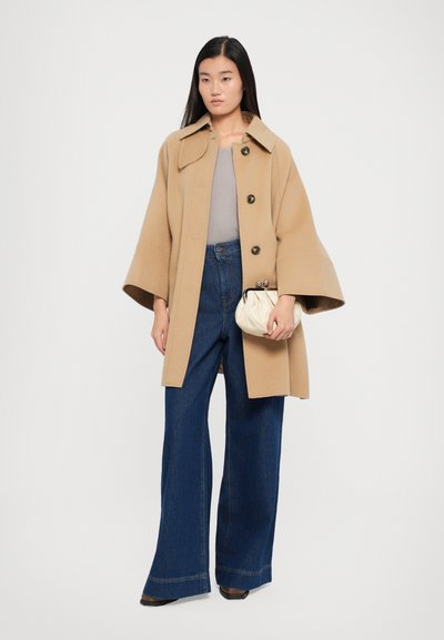 Vaaleanbeige villa-takki, jossa on leveät hihat ja suuret napit, yhdistettynä korkeavyötäröisiin tummansinisii leveisiin farkkuihin ja kermaiseen clutch-laukkuun.