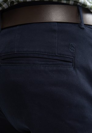 Donkerblauwe chino's met een gladde textuur, voorzien van een achterzak en geborduurd logo-detail. Een bruine leren riem maakt de outfit compleet.