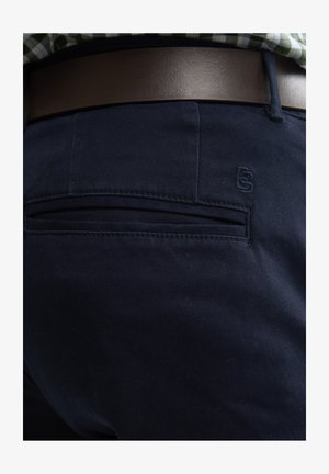 Chino marine foncé avec une texture lisse, doté d'une poche arrière et d'un détail de logo brodé. Une ceinture en cuir marron complète la tenue.