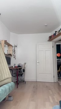 Soggiorno con un divano verde in tessuto, scrivania in legno e sedia da ufficio. Pavimento in legno chiaro, pareti bianche e una porta bianca chiusa.
