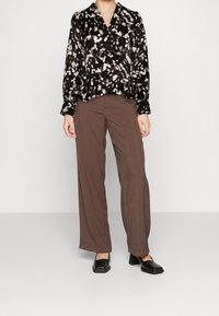 Blusa a fantasia nero e crema con design avvolgente, abbinata a pantaloni a gamba larga marroni a vita alta. Completata con scarpe nere dalla punta quadrata.