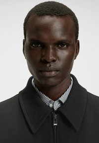 Veste zippée noire avec un col structuré, portée sur une chemise rayée noir et blanc. Tissu lisse avec une coupe ajustée.
