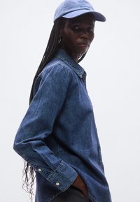 Tummansininen farkkupaita, jossa on napillinen kaulus ja pitkät hihat. Yhdistetty vaaleansiniseen lippalakkiin, jossa on ommeltu brändäys edessä.