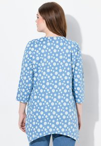 Lichtblauwe tuniek met witte stippen, driekwart mouwen, een hoge-lage zoom en een gladde katoentextuur. Eenvoudige, gebogen halslijn.