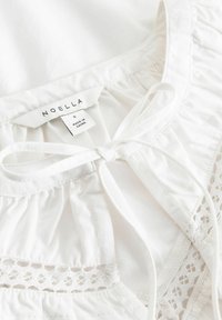 Biała bawełniana bluzka z falbanianym dekoltem, wyposażona w zapięcie na kokardę oraz koronkową lamówkę przy dole. Metka "NOELLA", rozmiar S, wyprodukowana w Chinach.