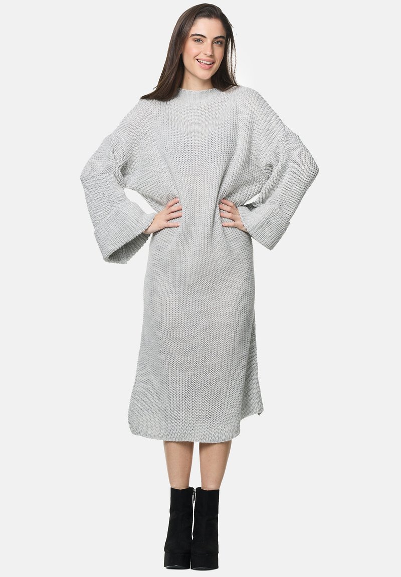 Helles graues gestricktes Kleid mit lockerem Schnitt, weitem Ärmeln und knielangem Saum. Getragen mit schwarzen Stiefeln bis zur Knöchel. Strukturierte Stoffdetails.