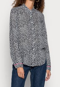 Nainen pukeutunut mustaan napitettuun paitaan, jossa on valkoinen pallokuosi ja vaaleanpunainen somiste kalvosimissa, yhdistettynä sinisiin farkkuihin.