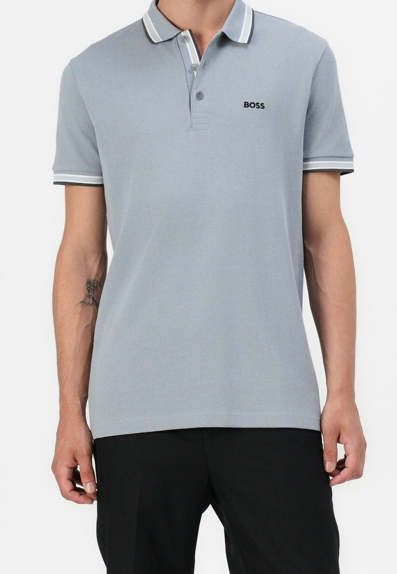 Homme portant un polo à manches courtes gris clair avec une bordure blanche et le logo "BOSS" sur la poitrine, associé à un pantalon noir.