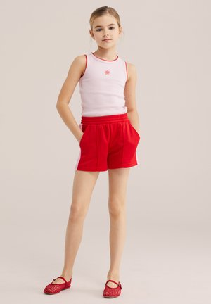 Jeune fille debout, mains dans les poches, portant un haut sans manches blanc avec bordure rouge, un short rouge et des chaussures plates rouges sur fond neutre.