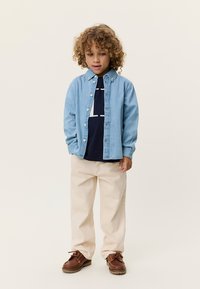 Ανοιχτό μπλε τζιν πουκάμισο, σκούρα μπλε μπλούζα με λευκή εκτύπωση, παντελόνι σε εκρού χρώμα και καφέ παπούτσια. Το σύνολο διαθέτει άνετη εφαρμογή και απαλότητα.