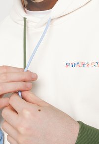 Kermansävyinen huppari, jossa vihreät ja vaaleansiniset nyörit. Hupussa kirjailtu värikäs "SCOTCH & SODA" -logo. Sileä kangaspinta.