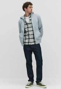 Jeune homme debout, les mains dans les poches, portant un sweat à capuche gris zippé, une chemise à carreaux verte et blanche, un jean sombre et des baskets vertes, regardant sur le côté.