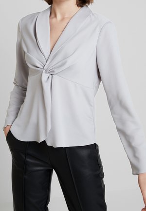 Kobieta ubrana w jasnoszarą bluzkę z długim rękawem z przednim skręceniem oraz czarne skórzane spodnie, stoi z jedną ręką w kieszeni.