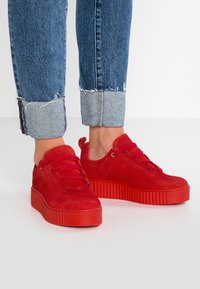 Baskets en daim rouge avec une semelle plate-forme texturée, dotées de lacets ronds et d'un design bas. Les jeans ont un ourlet effiloché et un revers.