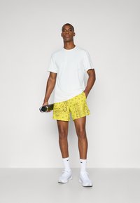 Camiseta blanca con pequeños agujeros, pantalones cortos amarillos brillantes con un diseño floral, y zapatillas deportivas blancas con detalles en negro.
