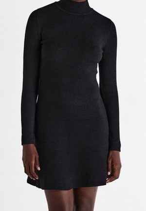 Kobieta ubrana w dopasowaną czarną mini sukienkę z długim rękawem i golfem na jednolitym jasnym tle.