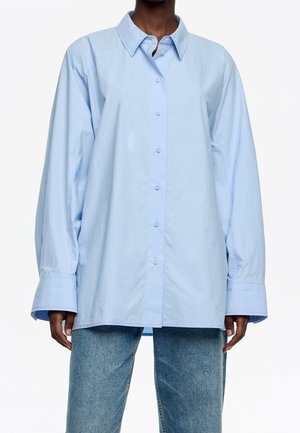 Personne portant une chemise ample bleu clair à boutons avec manches longues et un jean bleu, debout devant un fond blanc uni.