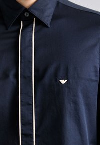 Marineblaues Hemd mit Knöpfen und kontrastierendem cremefarbenem Paspel entlang der Front sowie einem kleinen gestickten Logo auf der Brust. Glatte Stoffstruktur.