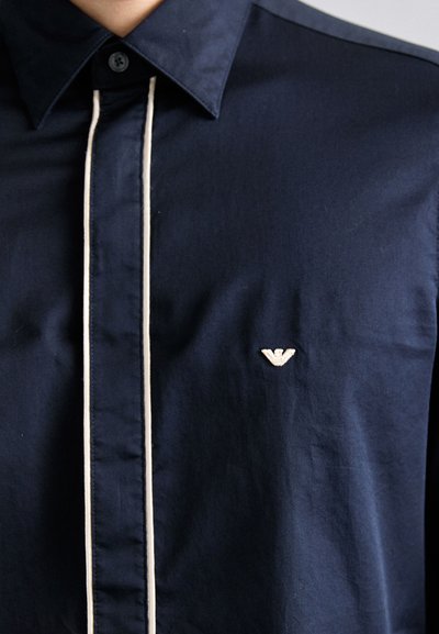 Mörkblå skjorta med knappar och kontrasterande krämfärgad piping fram samt en liten broderad logotyp på bröstet. Slät tygkvalitet.
