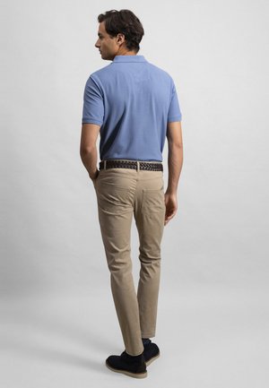 Mann trägt blaues Poloshirt, beigefarbene Hose mit geflochtenem Gürtel und schwarze Schuhe, steht mit dem Rücken zum Betrachter vor einem schlichten Hintergrund.