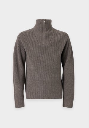 Jeune homme portant un pull gris à demi-fermeture éclair sur une chemise blanche, pantalon noir sur mesure et chaussures en cuir noires, debout avec une main dans la poche.
