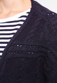 Stickad mörk marinblå kofta med en texturerad kabeldesign, öppen front och bred sjalkrage, lagd över en svart och vit randig topp.