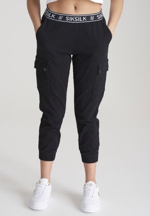 Femme portant un jogging cargo noir avec poignets élastiques et ceinture marquée Siksilk, associée à des baskets blanches sur un fond uni.