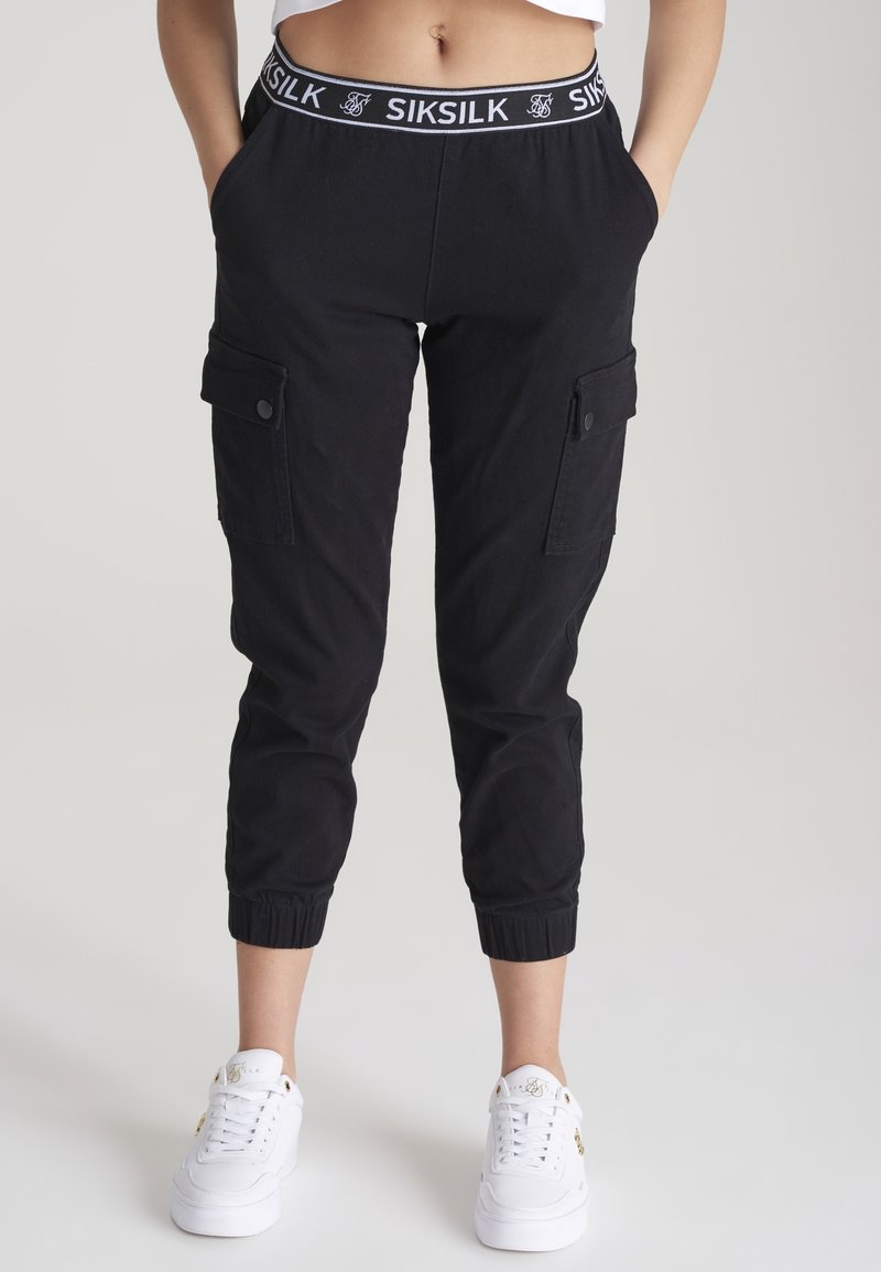 Femme portant un jogging cargo noir avec poignets élastiques et ceinture marquée Siksilk, associée à des baskets blanches sur un fond uni.