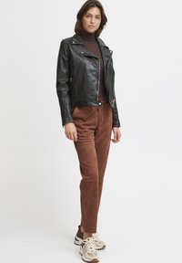 Black leather motorcycle jacket with zippered pockets, brown turtleneck, rust-colored suede pants, and beige sneakers with multicolored accents.