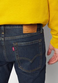 Vaqueros de mezclilla azul oscuro con costuras amarillas, etiqueta de cuero en la pretina, pestaña roja de Levi's en el bolsillo trasero, usados con una sudadera amarilla.