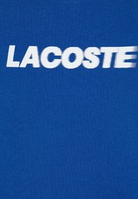 Blauwe stof met vetgedrukte witte tekst "LACOSTE" die iets vervaagd is aan de rechterkant.