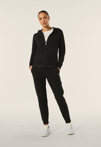 Felpa nera con zip e cappuccio, maniche lunghe e tasche frontali. Abbinata a pantaloni jogger neri con logo laterale e cinturino testurizzato.