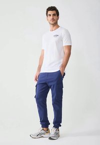 Muž stojí so založenými rukami v kapsách, oblečený v bielom tričku s krátkym rukávom, modrých cargo teplákoch a bielych behacích topánkach na jednoduchom pozadí.