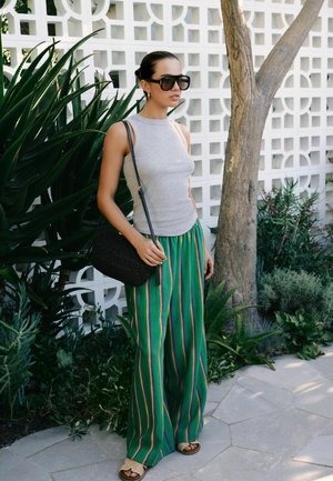 Moteris dėvi pilką rankovių neturintį viršutinį rūbą, žalias plačias dryžuotas kelnes, dideles saulės akinius ir neša juodą peties krepšį prie baltos raštuotos sienos ir augalų.