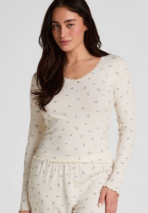 Jonge vrouw met lang donker haar, gekleed in een crème ribgebreide top met lange mouwen en bijpassende broek met een kleine blauwe bloemenprint en geschulpte randen.