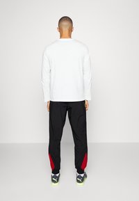 Weißes Langarmhemd und schwarze Hose mit roten Akzenten. Die Hose ist am Knöchel schmal zulaufend und hat einen Kordelzugbund. Sneakers sind schwarz mit Neonsohlen.
